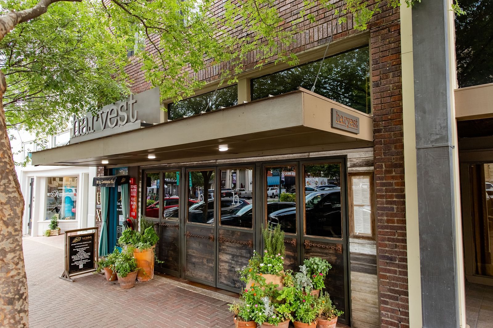 A restaurant exterior with large windows, a covered entrance, and potted plants along the sidewalk.- The Grand Hotel & Ballroom, McKinney