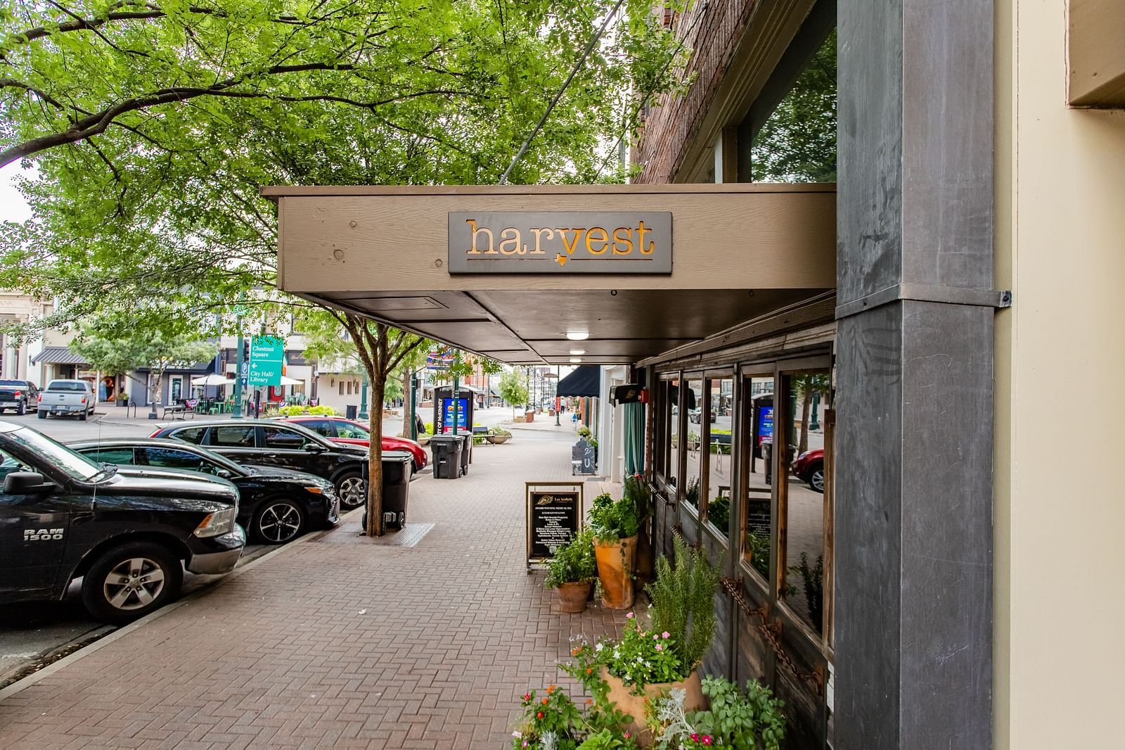 A storefront with a covered sidewalk features a  sign and lush greenery along the outdoor seating.- The Grand Hotel & Ballroom, McKinney
