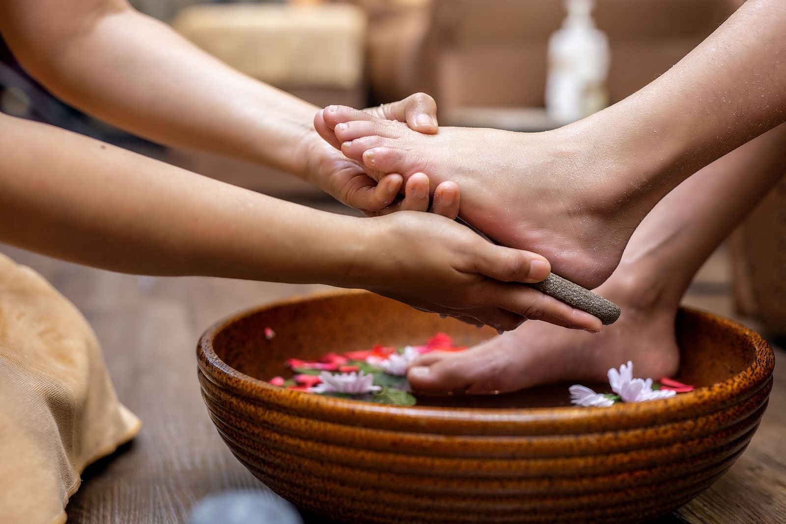Close-up of a foot spa treatment with a therapist gently massaging a person  feet over a wooden bowl filled with water and flower petals.