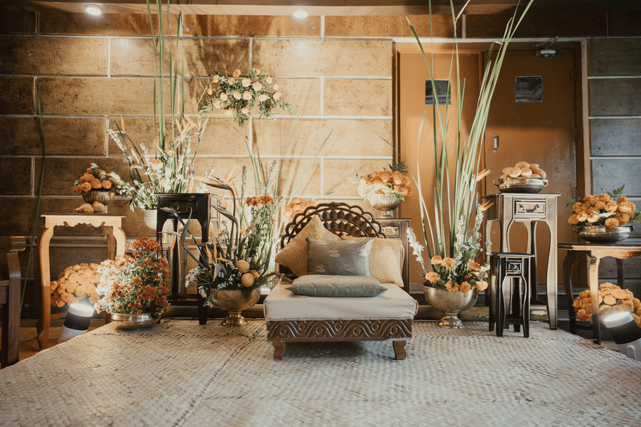 An indoor lounge area with a rustic aesthetic, featuring a light-colored sofa with patterned cushions, surrounded by various floral arrangements, decorative furniture, and tall plants against a textured wall.