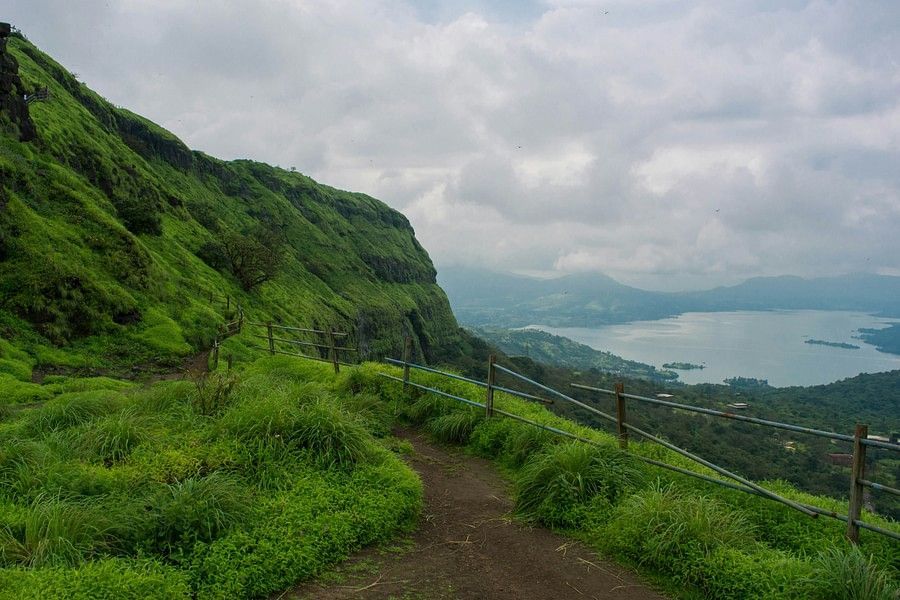 alt-text Lush green hillside with a narrow path curving along the slope, overlooking a distant lake and cloudy sky, conveying a sense of tranquil exploration.