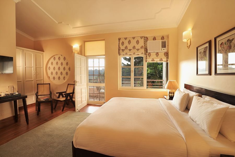 alt-text An image of a Super Deluxe Room featuring a double size bed, two chairs, a table where coffee maker is kept, and an open door from where sunlight is flooding the room - Lamrin Boutique Cottages Rishikesh