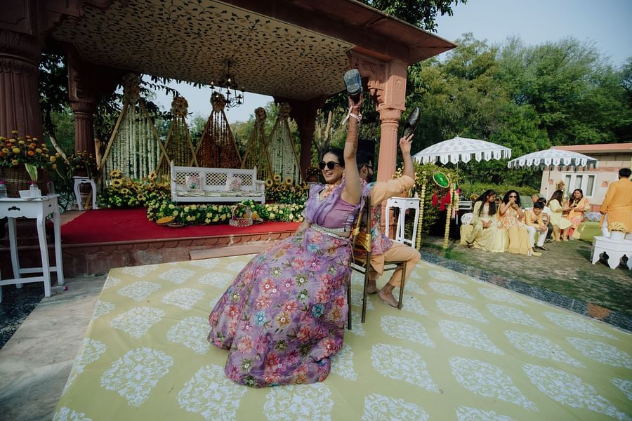 Bride seated and raising her arms while getting henna applied during the mehndi ceremony.