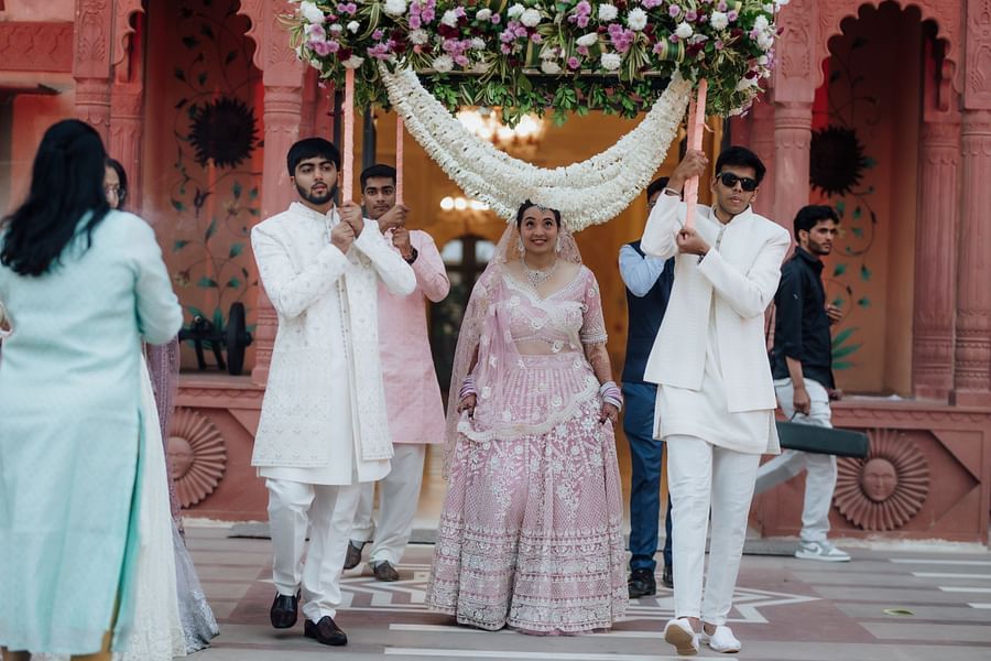 Bride in a pastel lehenga walking under a floral canopy with family members dressed in white.