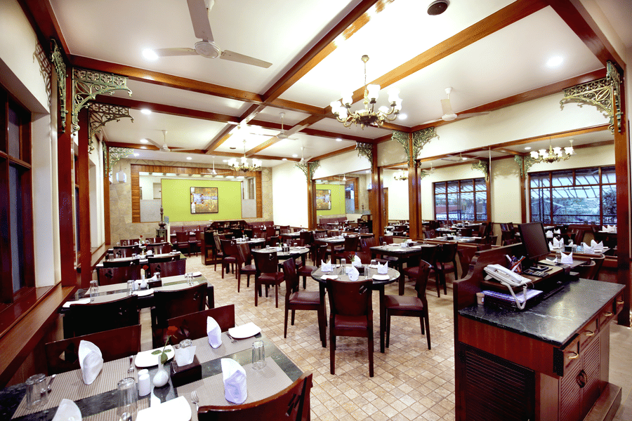 Spacious restaurant at Zara's Resort, Khandala, featuring a well-lit dining hall with neatly set tables and a chandelier, creating a warm, and inviting space for meals.