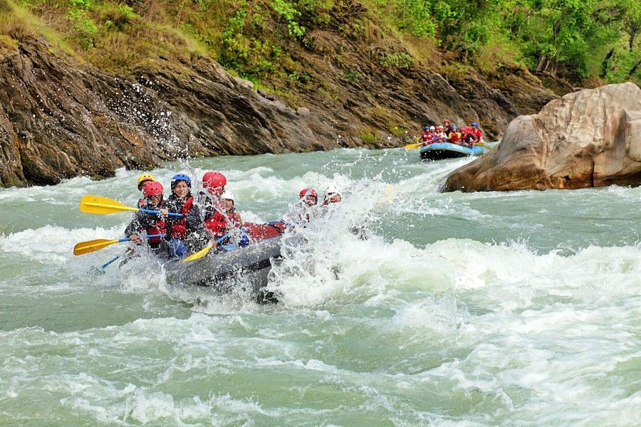 A group of people wearing red life jackets and helmets rafting down a fast-moving river surrounded by lush, green hills.