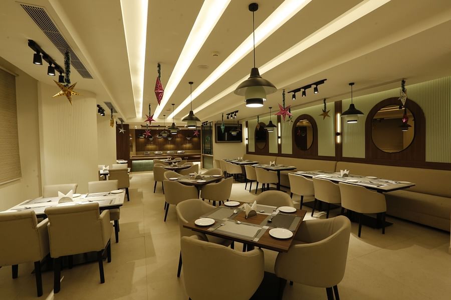 A poster of a restaurant at Tulip Inn, Zirakpur featuring several sets of tables, chairs, and sofa, lights hanging from the ceiling and mirrors attached on the wall