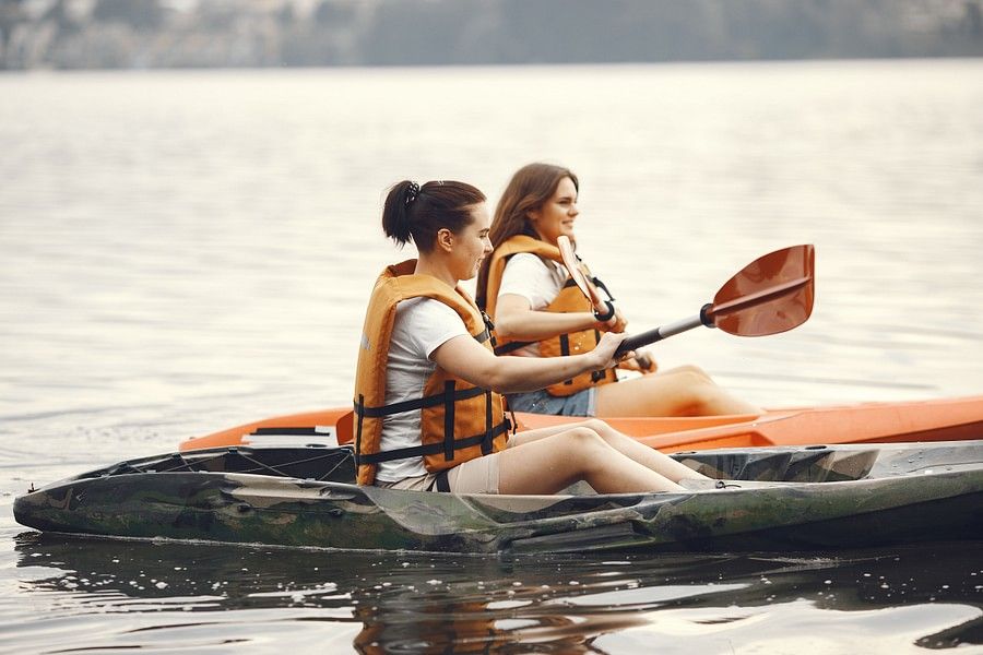 alt-text Two women in life jackets kayaking on a calm lake, surrounded by serene water and lush greenery.