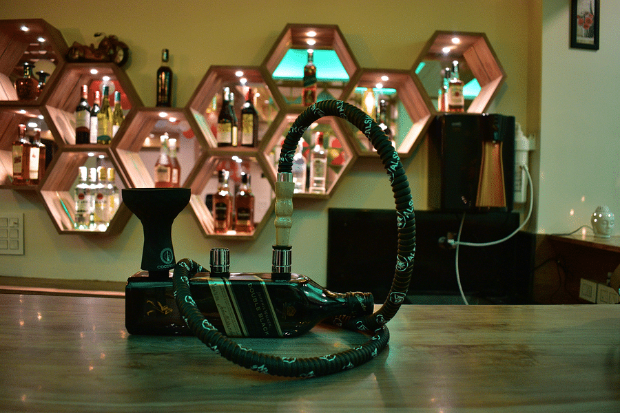 alt-text bar counter table showcasing a hookah made out of a bottle of alcohol on table and a backdrop of alcohol bottles kept in hexagonal boxes - Hotel Montana Vyoo