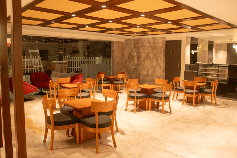 A hotel lobby area with wooden tables and chairs,  cushioned chairs, and ceiling lights arranged in a grid pattern.