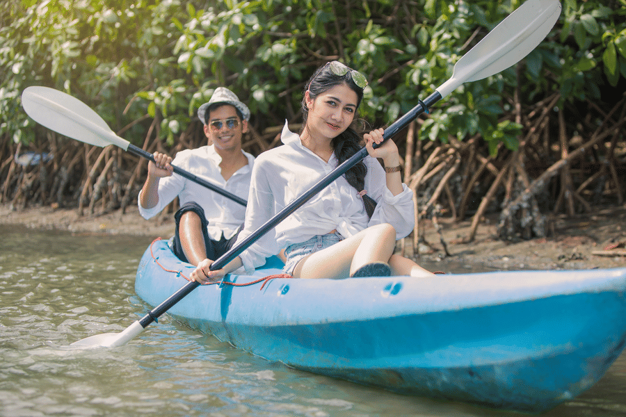 2 individuals, each in a kayak, paddling through calm river waters surrounded by dense mangroves, showing a peaceful and serene exploration setting.