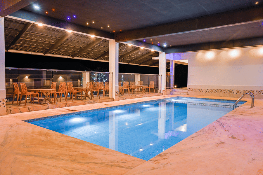 An outdoor swimming pool illuminated at night with several dining tables and chairs arranged on the surrounding deck at Abad Silvermist, Vagamon.