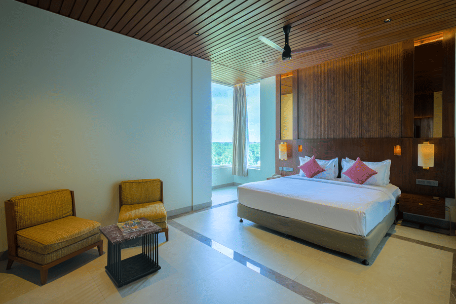 Hotel bedroom featuring a king size bed, two lounge chairs, wooden ceiling, warm lighting and a dark wood feature wall.