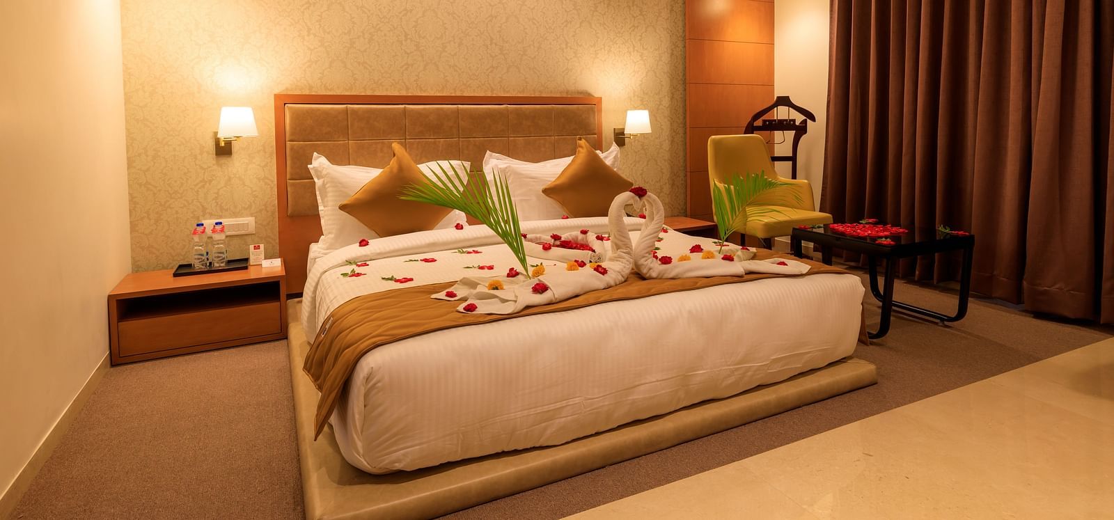A warmly lit hotel room at Lords Eco Inn Apple Gate features a decorated double bed, bedside tables, a chair, and brown curtains.