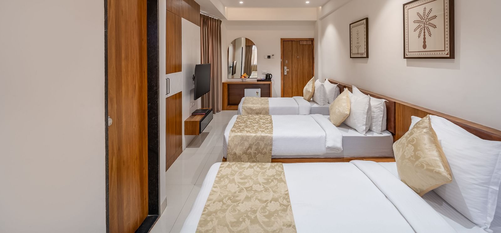 A hotel room features three single beds with white linens and decorative pillows, along with a TV and a vanity area - Lords Eco Inn Ambaji.