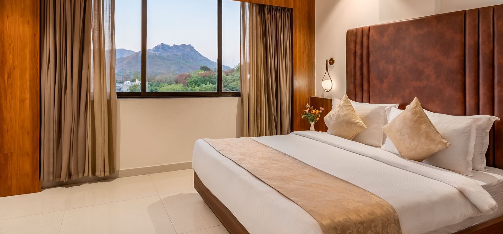 A comfortable king-size bed with soft pillows and a throw faces a window with curtains revealing a mountain view - Lords Eco Inn Ambaji.