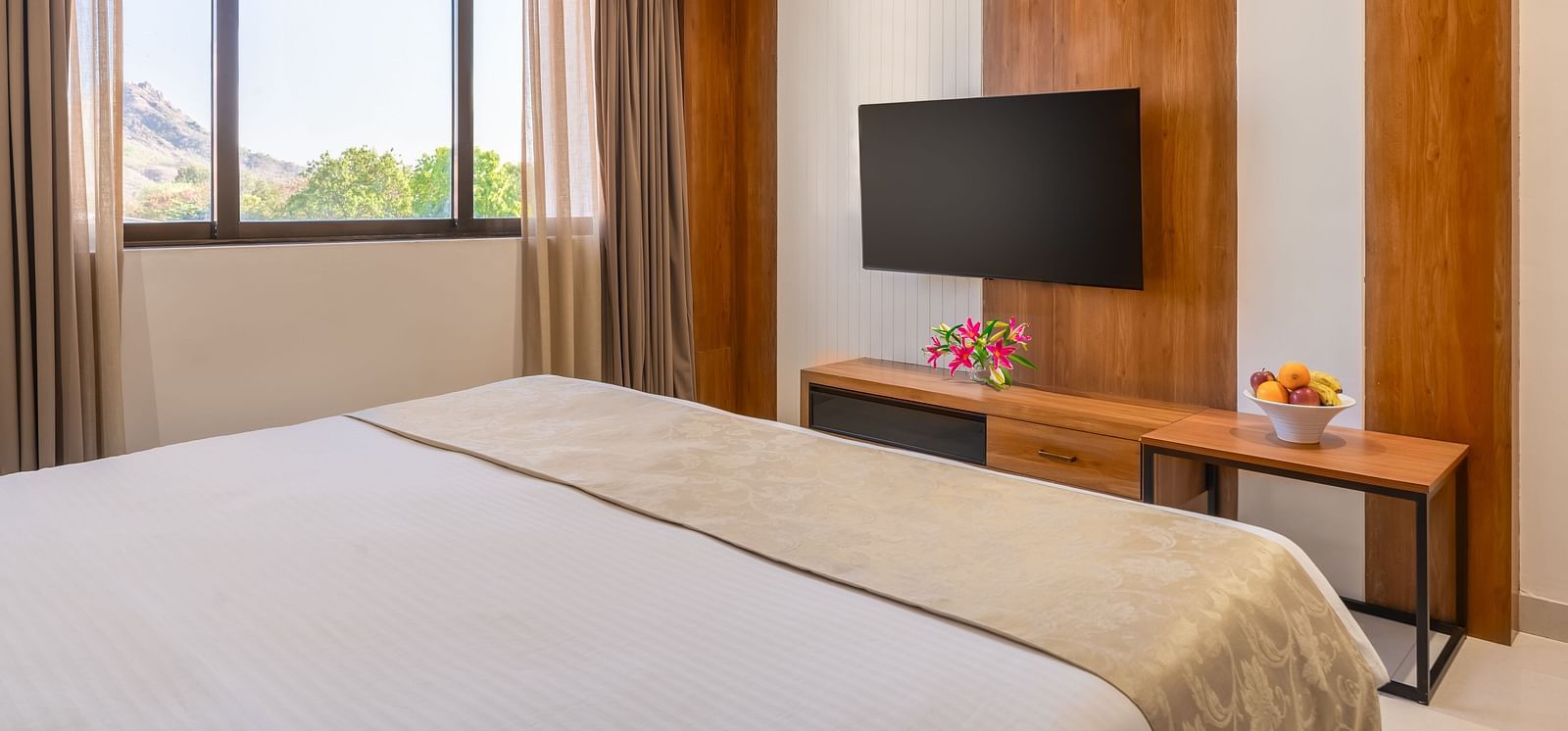 A comfortable bed with white linens and a decorative throw faces a mounted television in a well-lit hotel room with a view - Lords Eco Inn Ambaji.