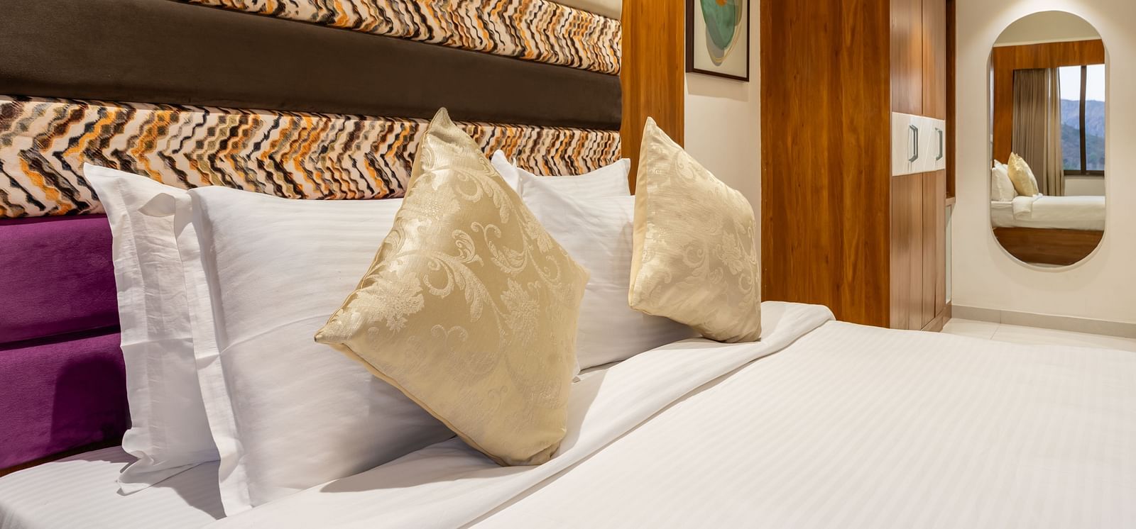  A bed with white linens and decorative pillows is set against a multi-textured headboard in a hotel room - Lords Eco Inn Ambaji.