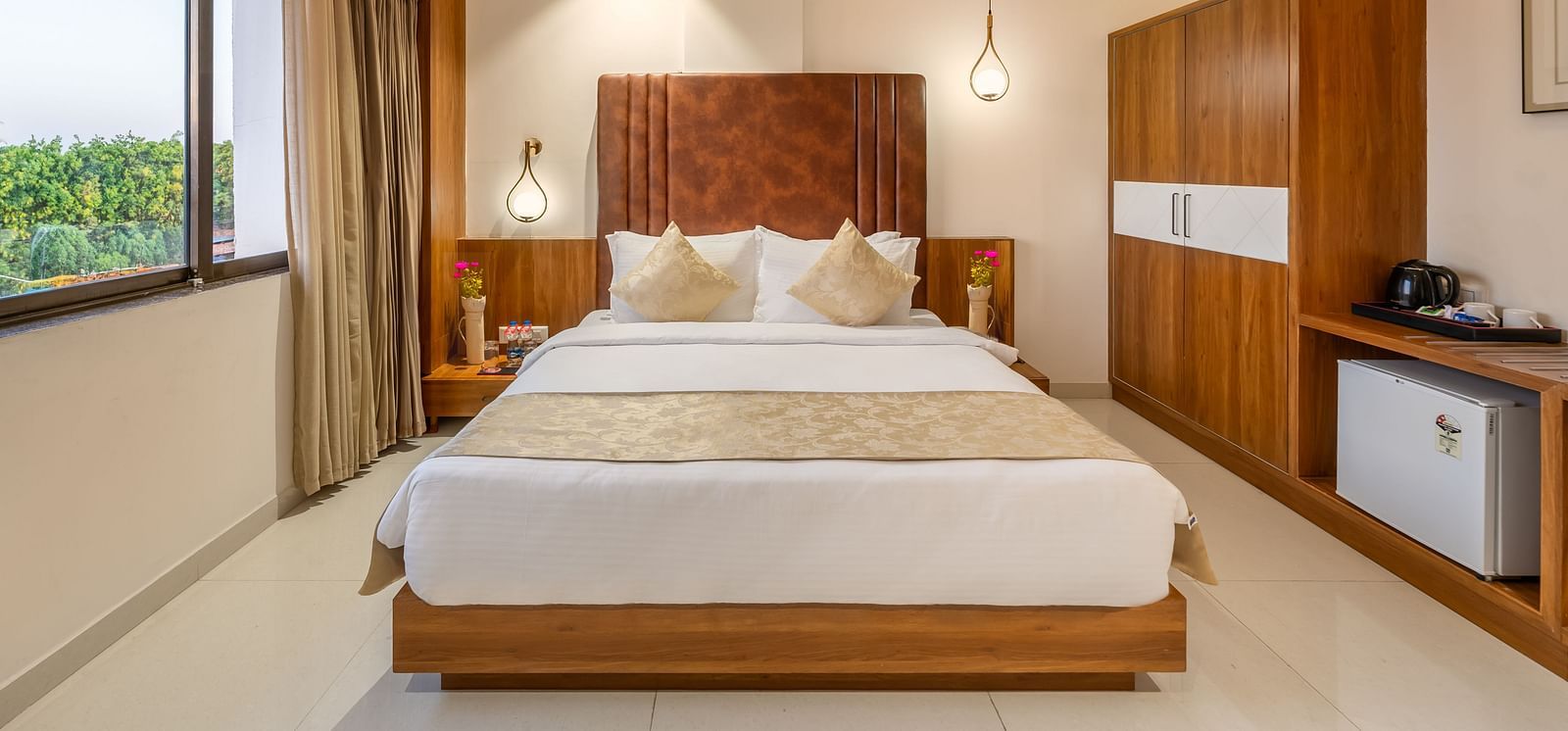 A comfortable king-size bed with white linens and decorative pillows is the focal point of a well-lit hotel room featuring a large window, a wooden wardrobe, and a refreshment station - Lords Eco Inn Ambaji.