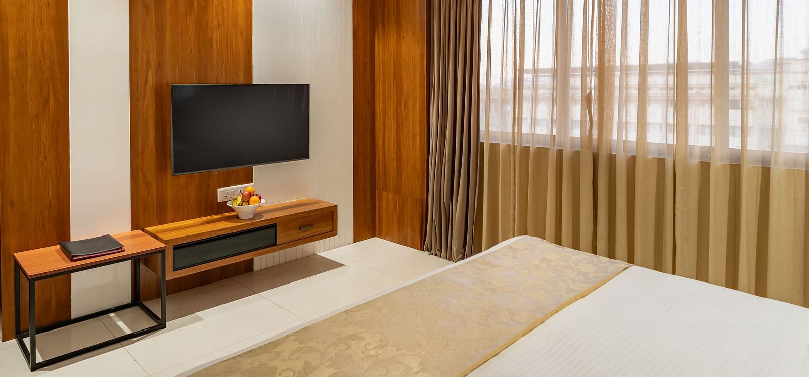 A comfortable bed with a decorative throw is situated in a hotel room with a mounted television and a window with layered curtains - Lords Eco Inn Ambaji.
