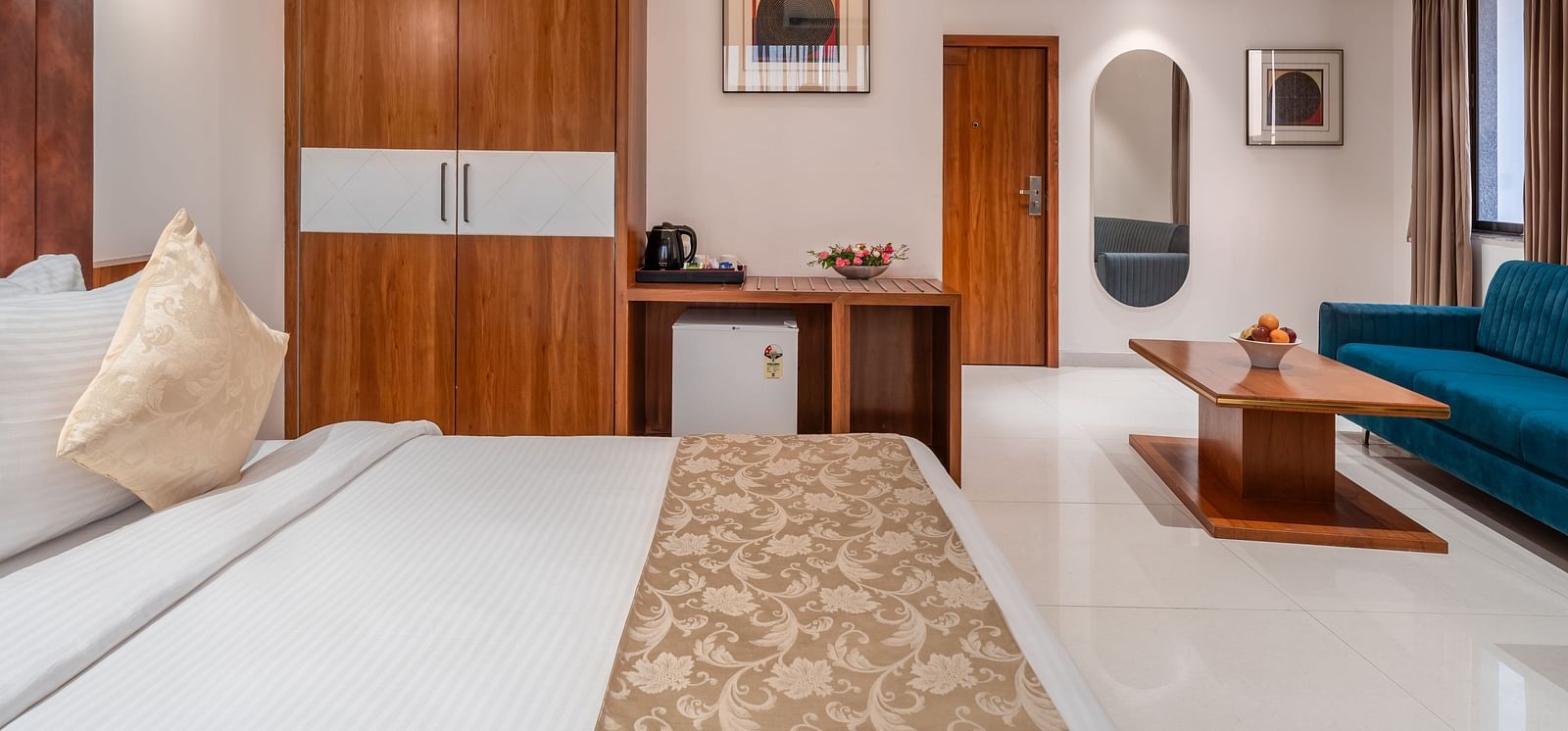 A comfortable bed faces a living area with a blue sofa and a wooden table in a well-lit hotel suite - Lords Eco Inn Ambaji.