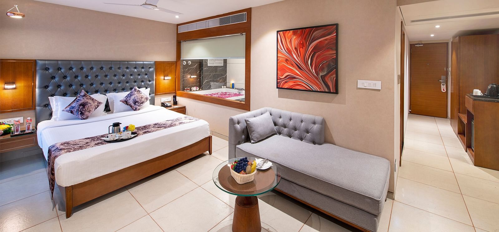 A spacious hotel room  featuring a large double bed with decorative pillows, a grey chaise lounge, and modern artwork on the wall -  SK Lords Inn Ahmedabad