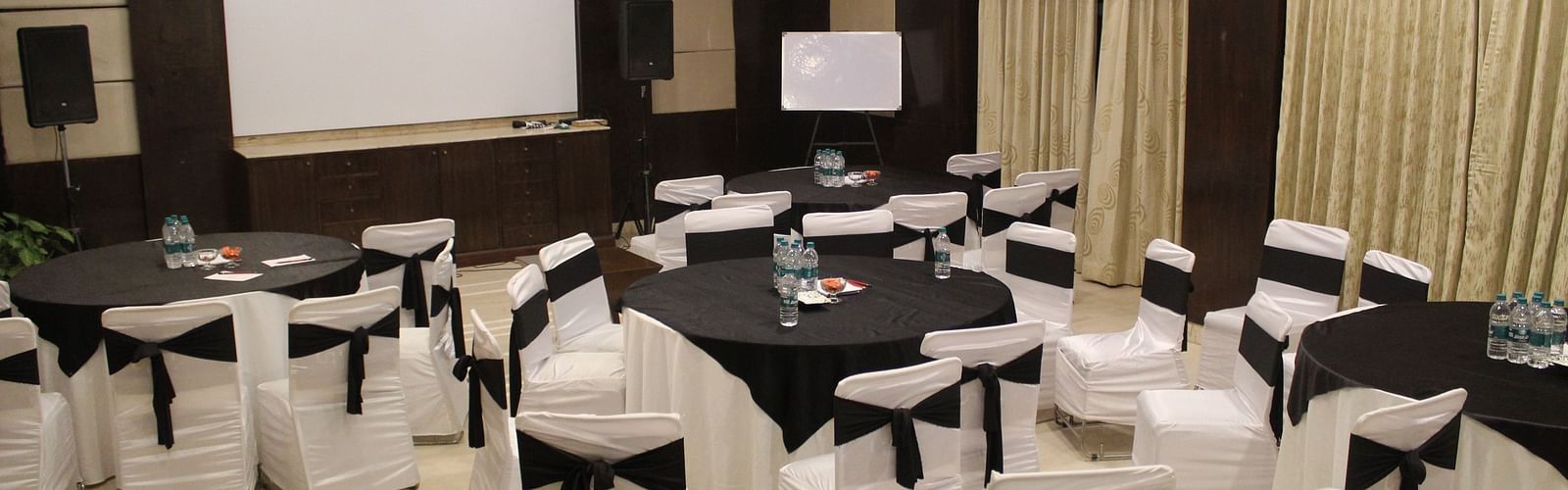 An image of a banquet hall at Inde Hotel Signature Tower, Gurgaon featuring a few round tables, each surrounded by chairs