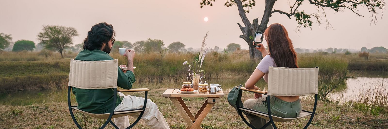 Two people sitting on chairs with a dining setup in an open grassland surrounded by trees at Trees N Tigers, Tadoba.
