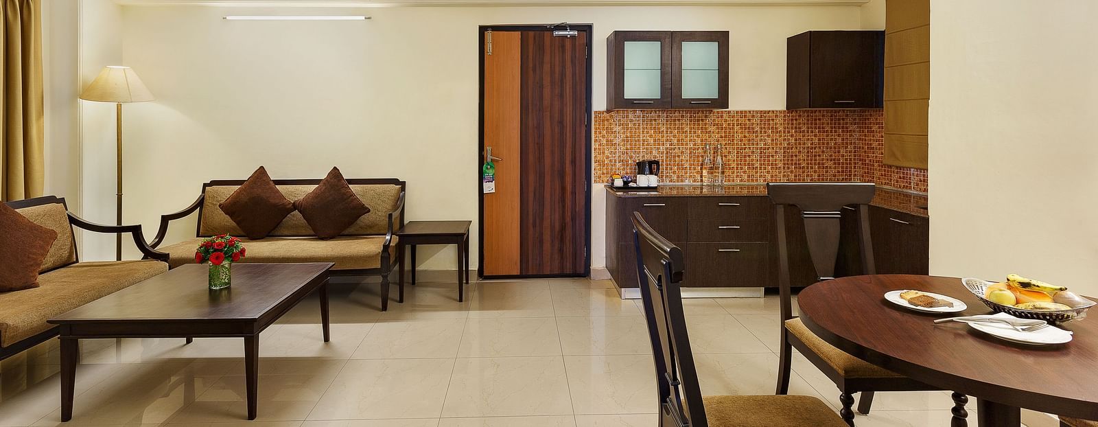 An image of a spacious and modern hotel suite. The living area features a comfortable sofa with throw pillows, a coffee table, and a floor lamp. A separate dining area