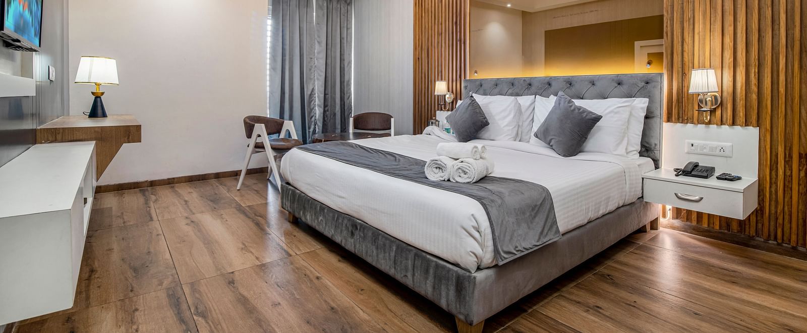 An image of the deluxe room featuring a bed, bed side tables, wall mounted flat screen TV, chairs and table, lamps and other room amenities - DLS Ganga Bliss, Haridwar