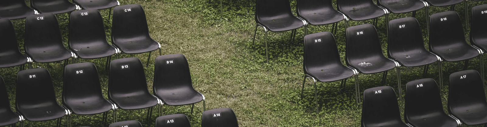 Rows of empty, dark plastic chairs set up in an open-air venue on a slightly overgrown green grass lawn.