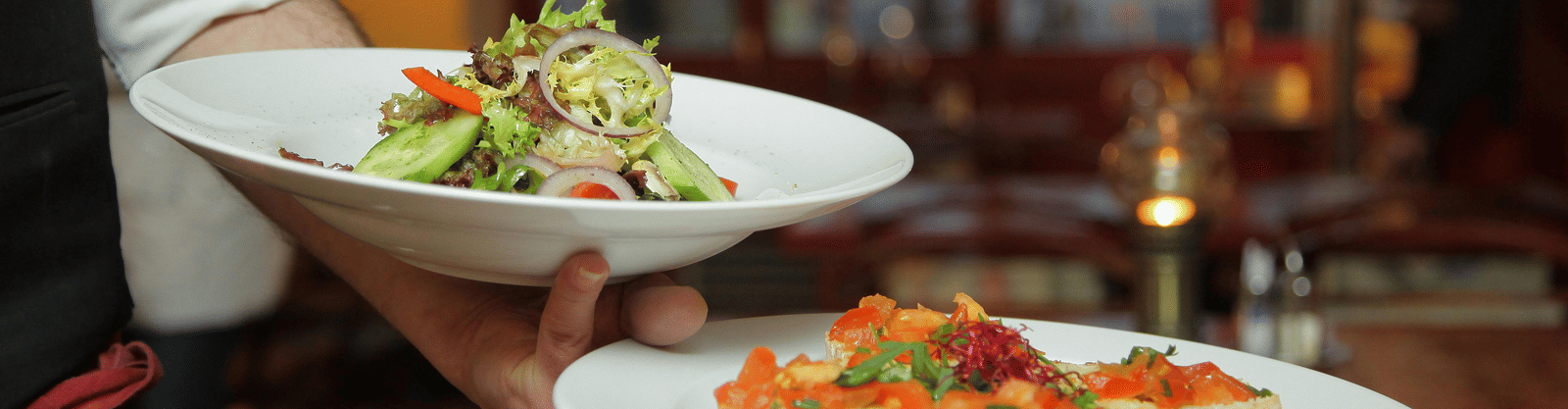 A waiter in a white shirt and black waistcoat is serving two plates of food, one with a side salad and one with bruschetta.