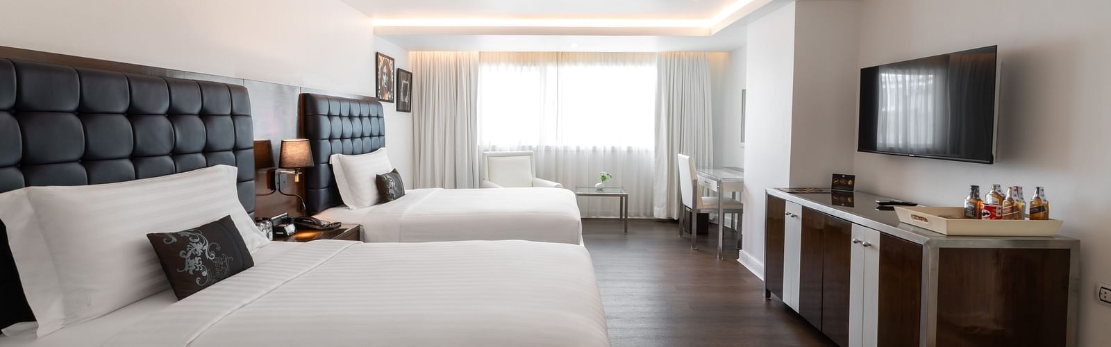 Modern hotel room with two large beds, white linens, dark headboards, a flat-screen TV, minibar, and a window letting in natural light at the far end of the room at Night Hotel Bangkok.