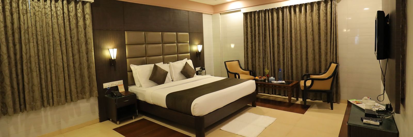 A front angle view of a bedroom at Krushnai Resort with a king size bed, a dark headboard, and brown drapes.