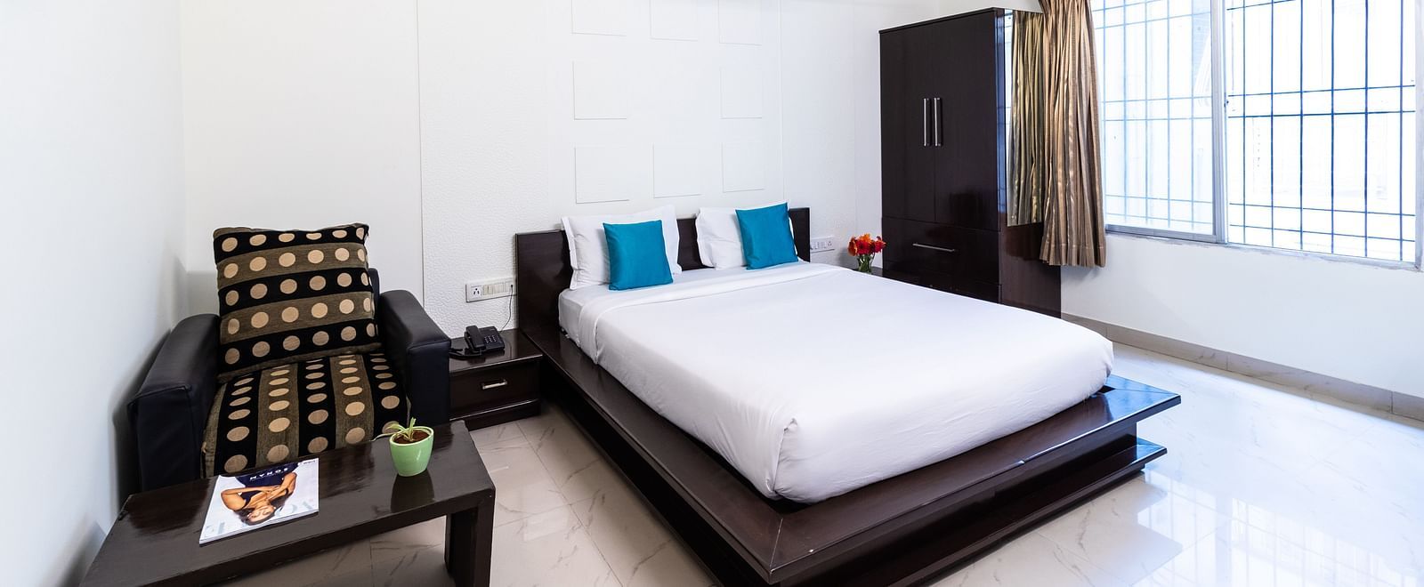 A room with a bed, an armchair, a wardrobe, and a large window at Sanctum Suites Domlur, Bangalore.