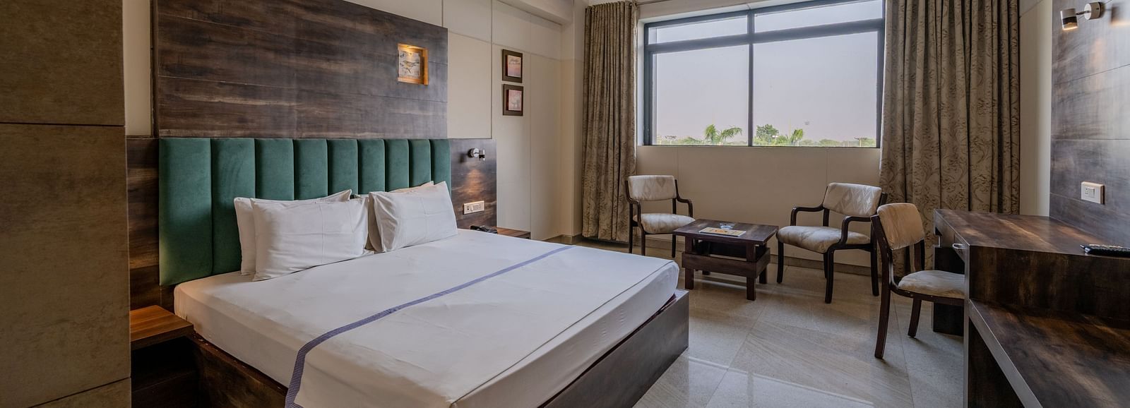 An accommodation in Hotel Highway King, Behror, featuring a plush double bed with upholstered headboard, seating area for two, a work desk and glass windows with open curtains.