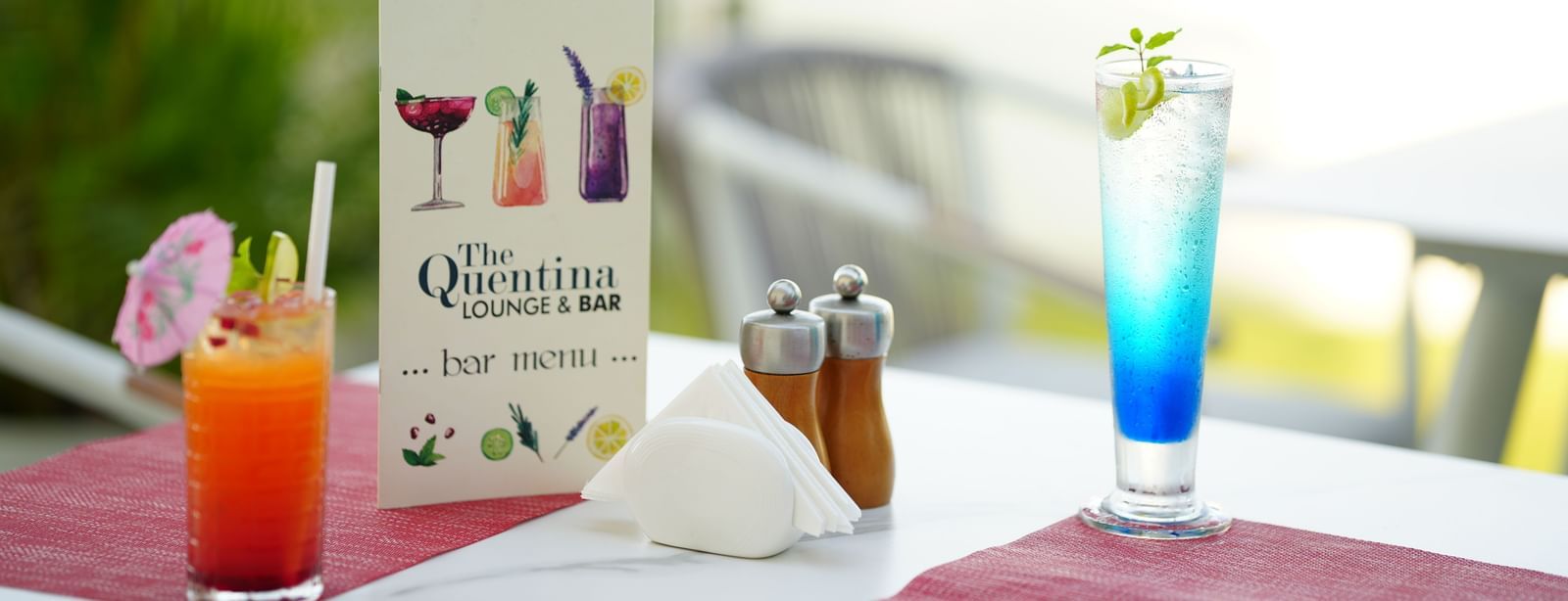 A red and a blue cocktail placed on an outdoor dining table with a menu card at Grande Bay Resort & Spa.