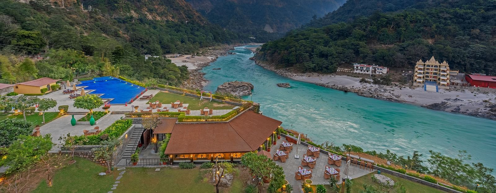 A panoramic view of Aloha on the Ganges, Rishikesh. The image shows the resort nestled amidst hills, with the Ganges River flowing peacefully in the foreground. There is a swimming pool, restaurant, and other facilities visible.