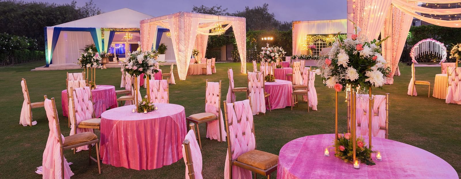 clusters of chairs and tables with pink covers and canopies lit up with decorative lights during the evening - The Hideaway Banquets & Lawns, Greater Noida