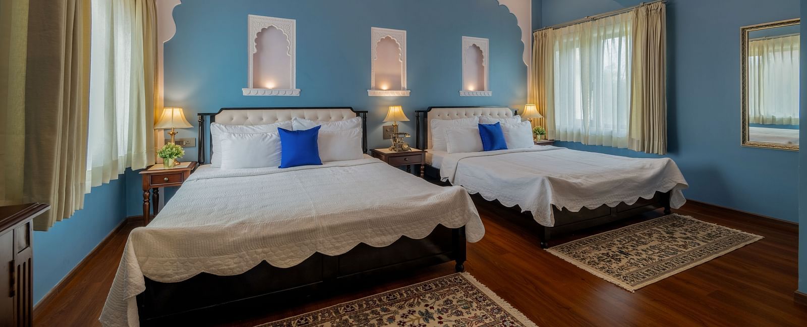 A blue-walled bedroom with two twin beds, blue cushions, arched niches, ceiling fans, and sheer curtains | Dera Rawatsar, Jaipur