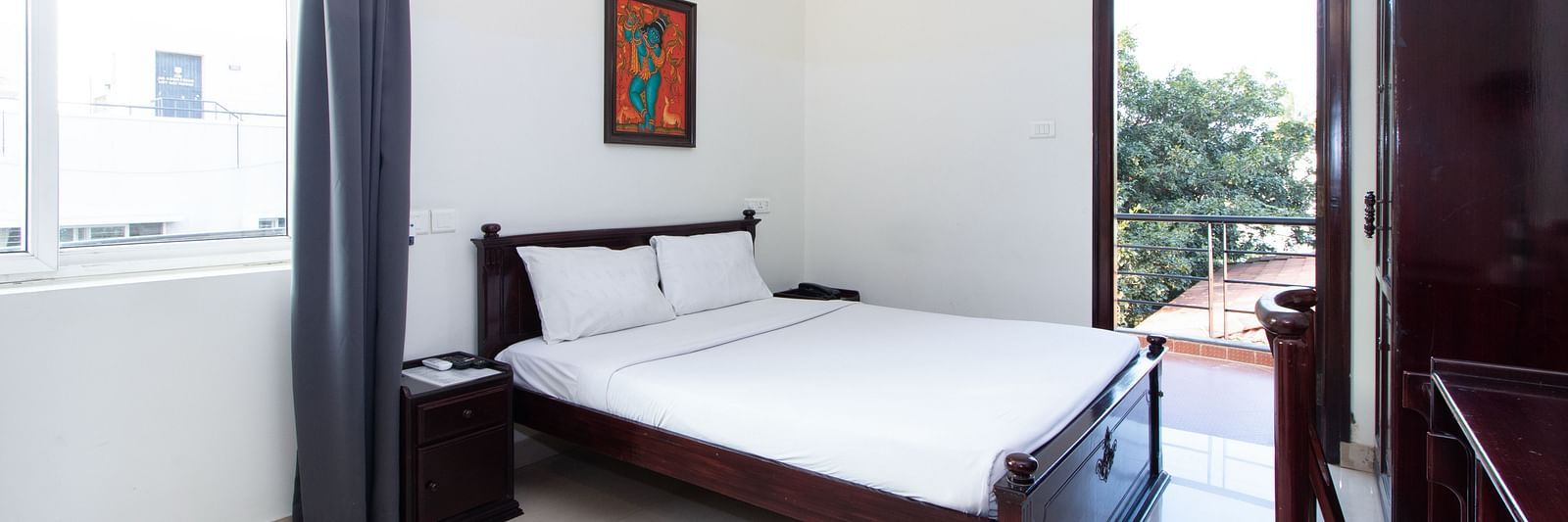 A room displaying a double bed with white bedding, a bedside table with a lamp, a wardrobe, a doorway leading to the balcony at Sanctum Suites Indiranagar, Bangalore.