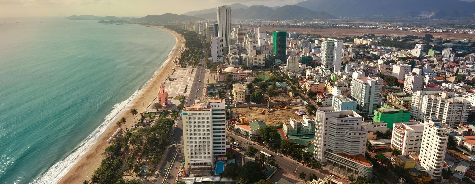 Aerial view of a coastal city, showcasing a long beach, buildings, and distant mountains under a partly cloudy sky.