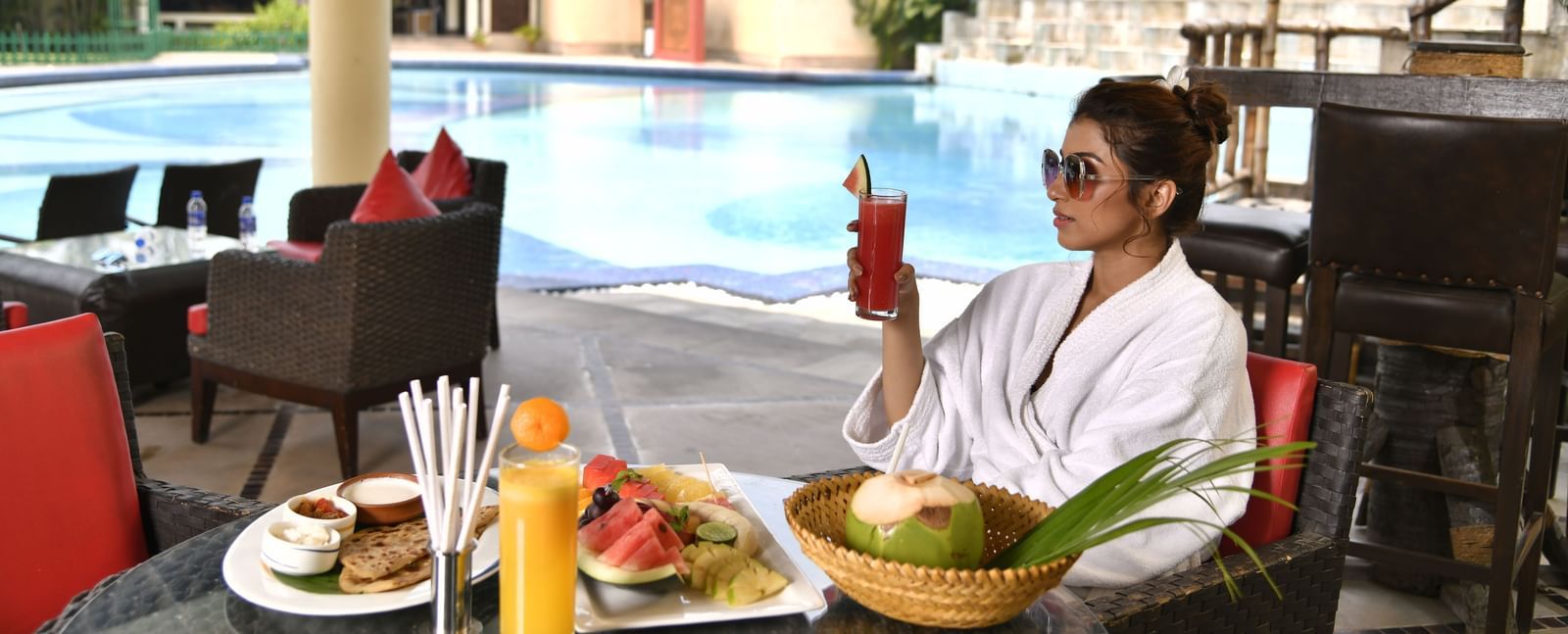 A woman in a white robe sitting by the pool, holding a drink in her hand, with a round wooden table beside her featuring several served dishes.