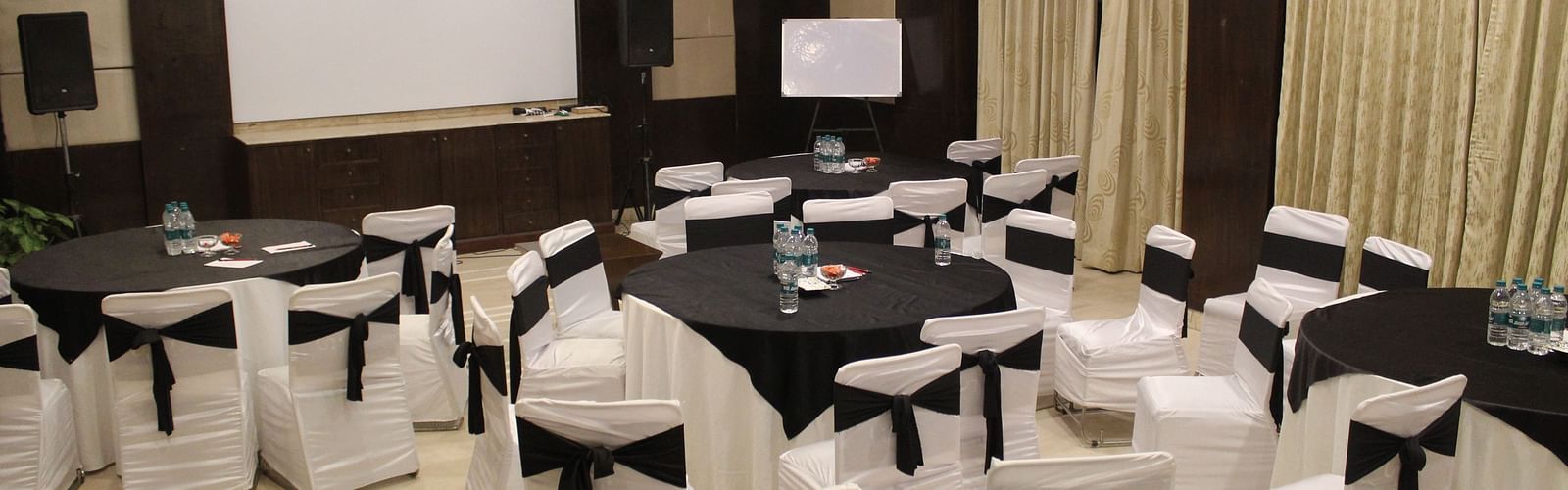 An image of a banquet hall at Inde Hotel Signature Tower, Gurgaon featuring a few round tables, each surrounded by chairs