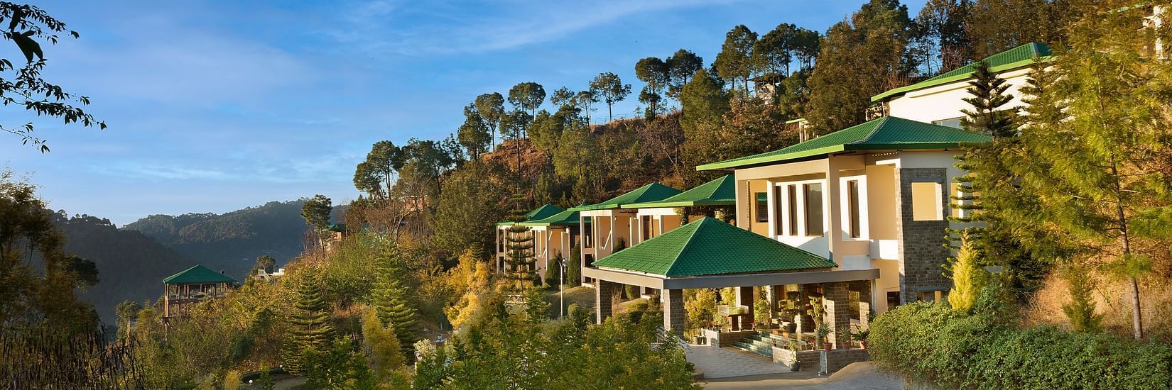 Scenic view of a hillside property surrounded by lush greenery and forested hills under a clear blue sky - Suryavilas Luxury Resort and Spa.