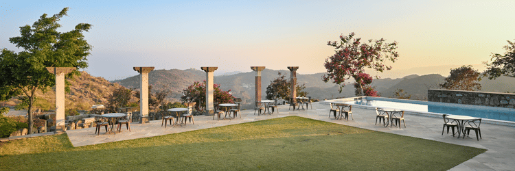 Lawn at Fateh Safari Suites, Kumbhalgarh featuring trimmed grass, surrounded by chairs and tables and a pool nearby with a view of green hills.