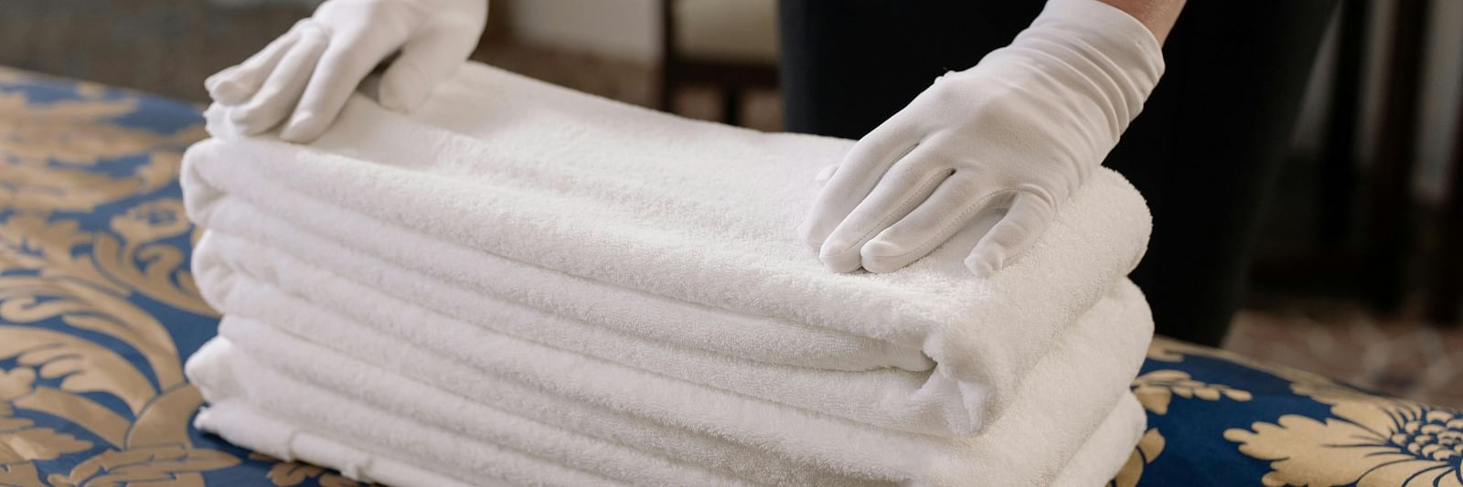 Hands in white gloves on neatly stack folded white towels on a bed with a blue and gold patterned spread.