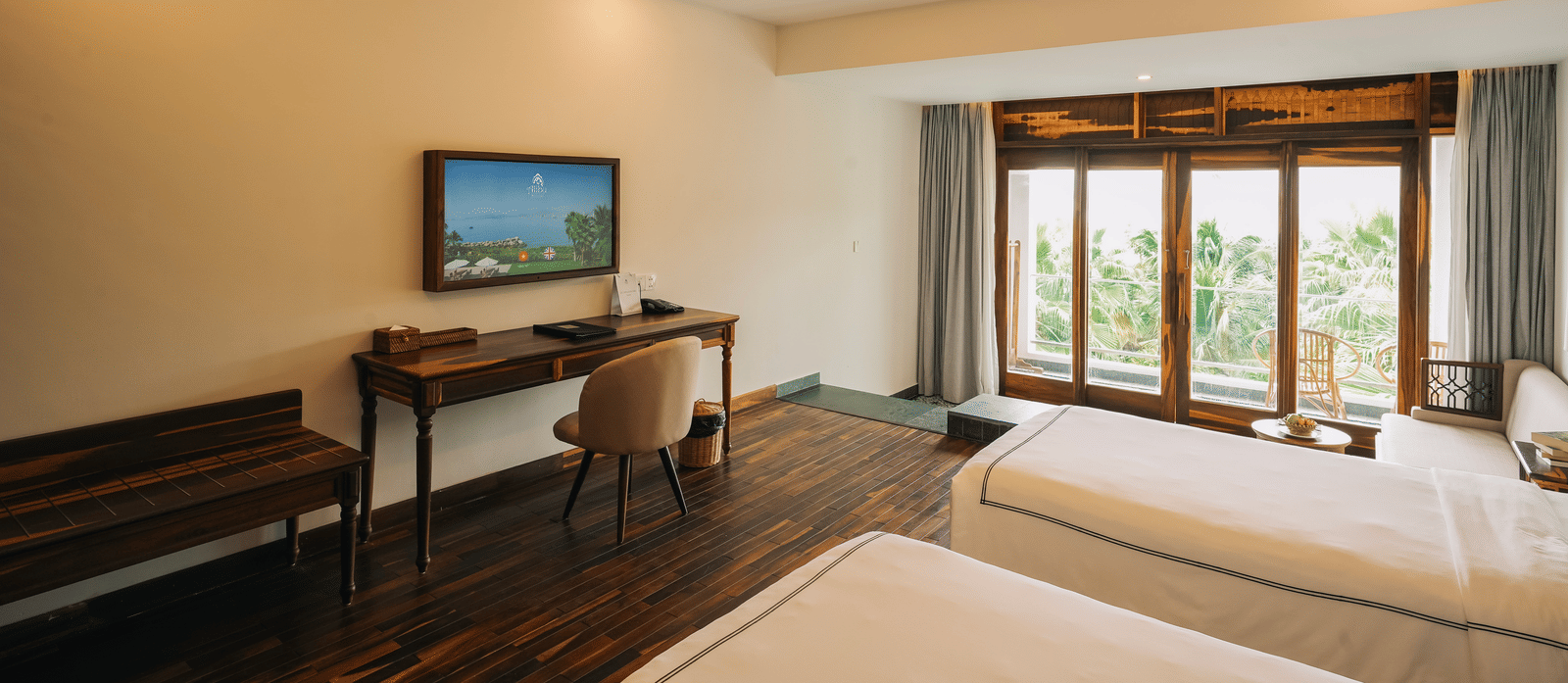 A bed at Alibu Resort Nha Trang featuring twin bed, a TV attached on the wall, a table, and windows with curtains.