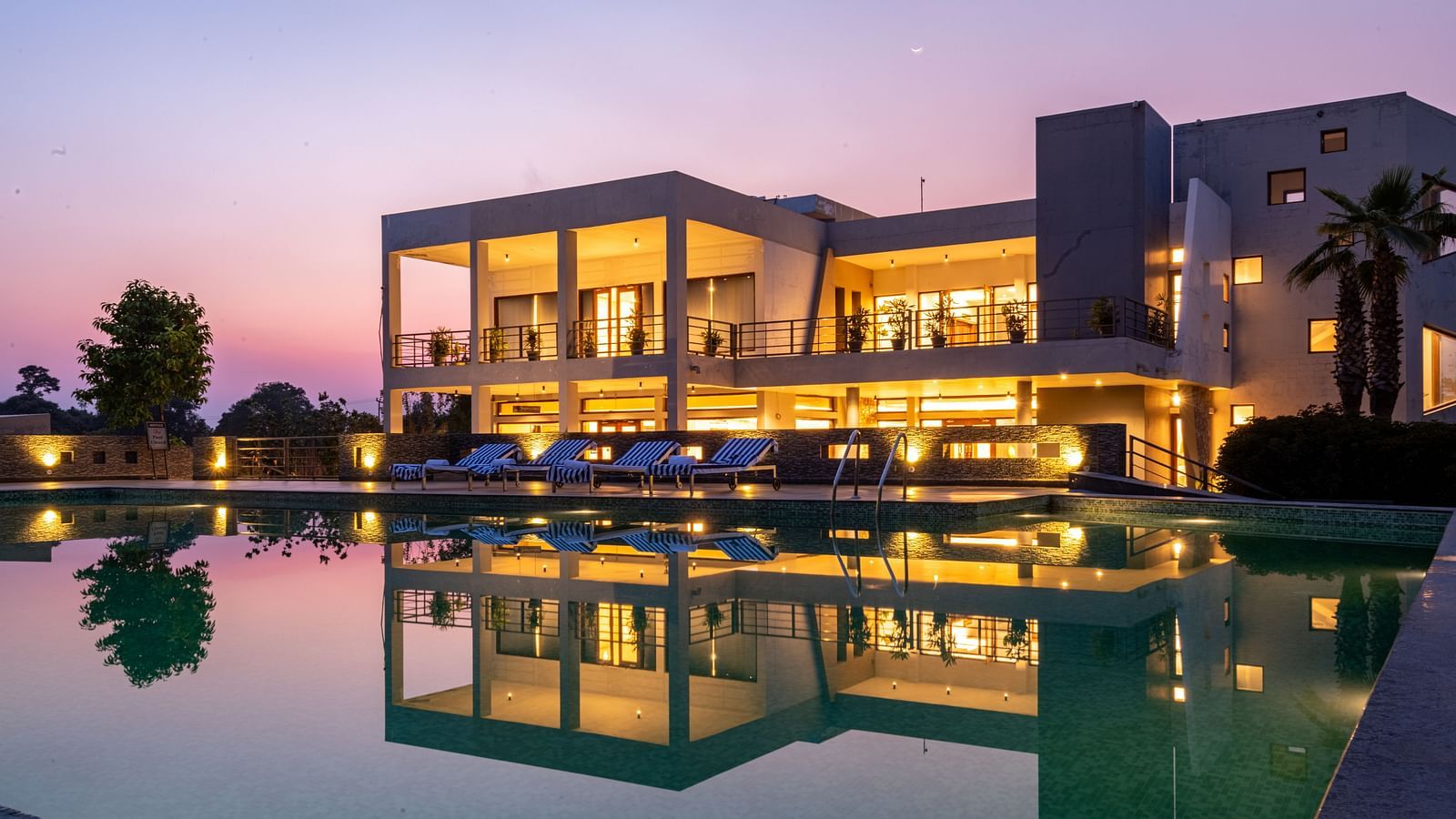Facade view of DLS Forest Vines Resort, with warm light glowing from its windows and doors, reflected on the shimmering swimming pool beneath a clear sky.