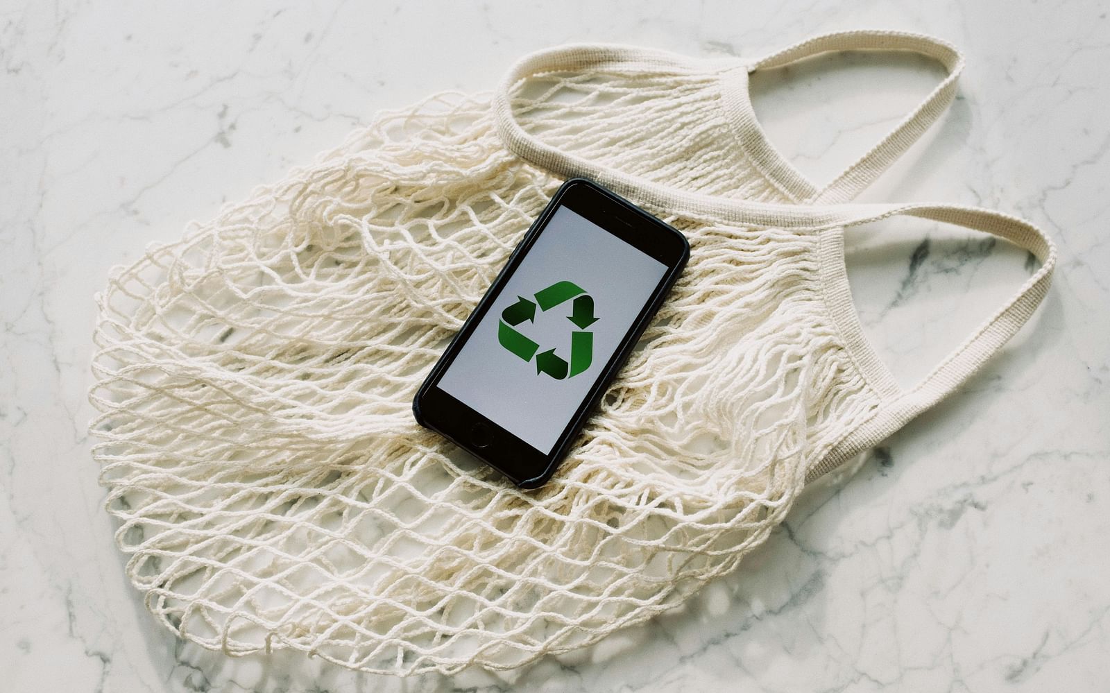 A white mesh reusable shopping bag with a smartphone resting inside it. The phone's screen displays a green recycling symbol.